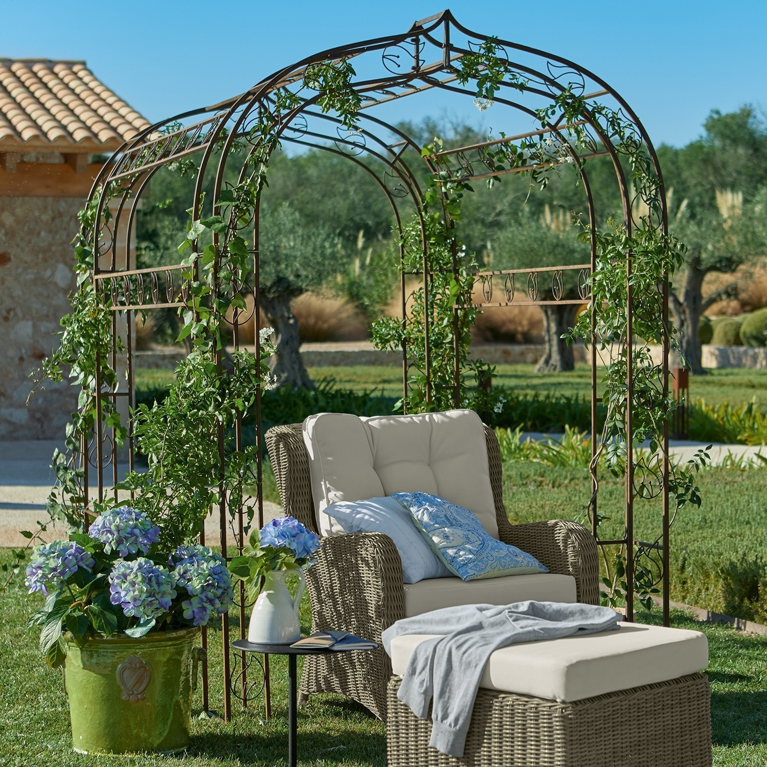 Metall-Gartenpergola mit grünen Kletterpflanzen und beigem Rattansessel mit Hocker