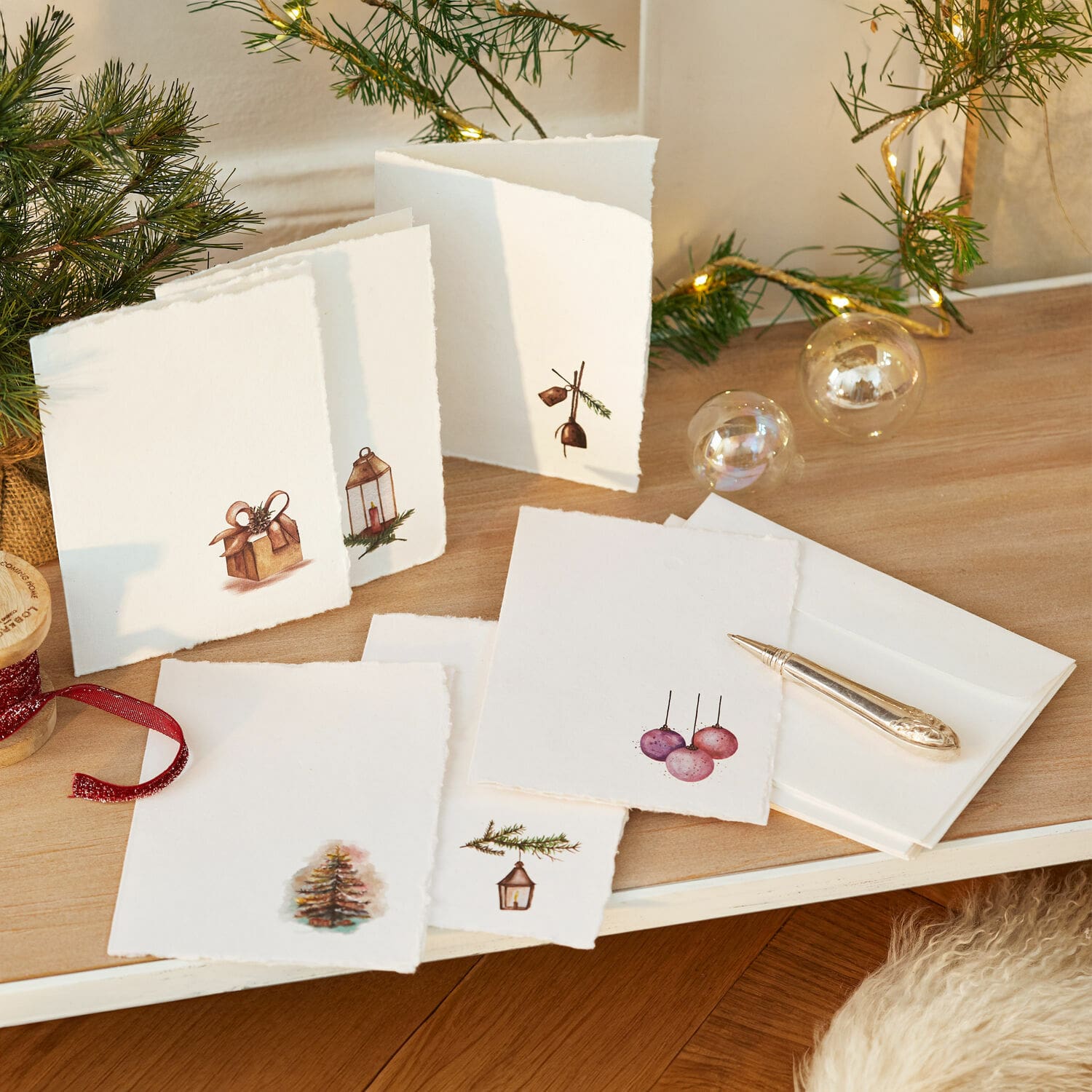 Grußkarten 6er Set Aeloria Set aus weißen Weihnachtskarten mit Tannen- und Geschenk-Motiven auf Holztisch mit LED-Lichterkette