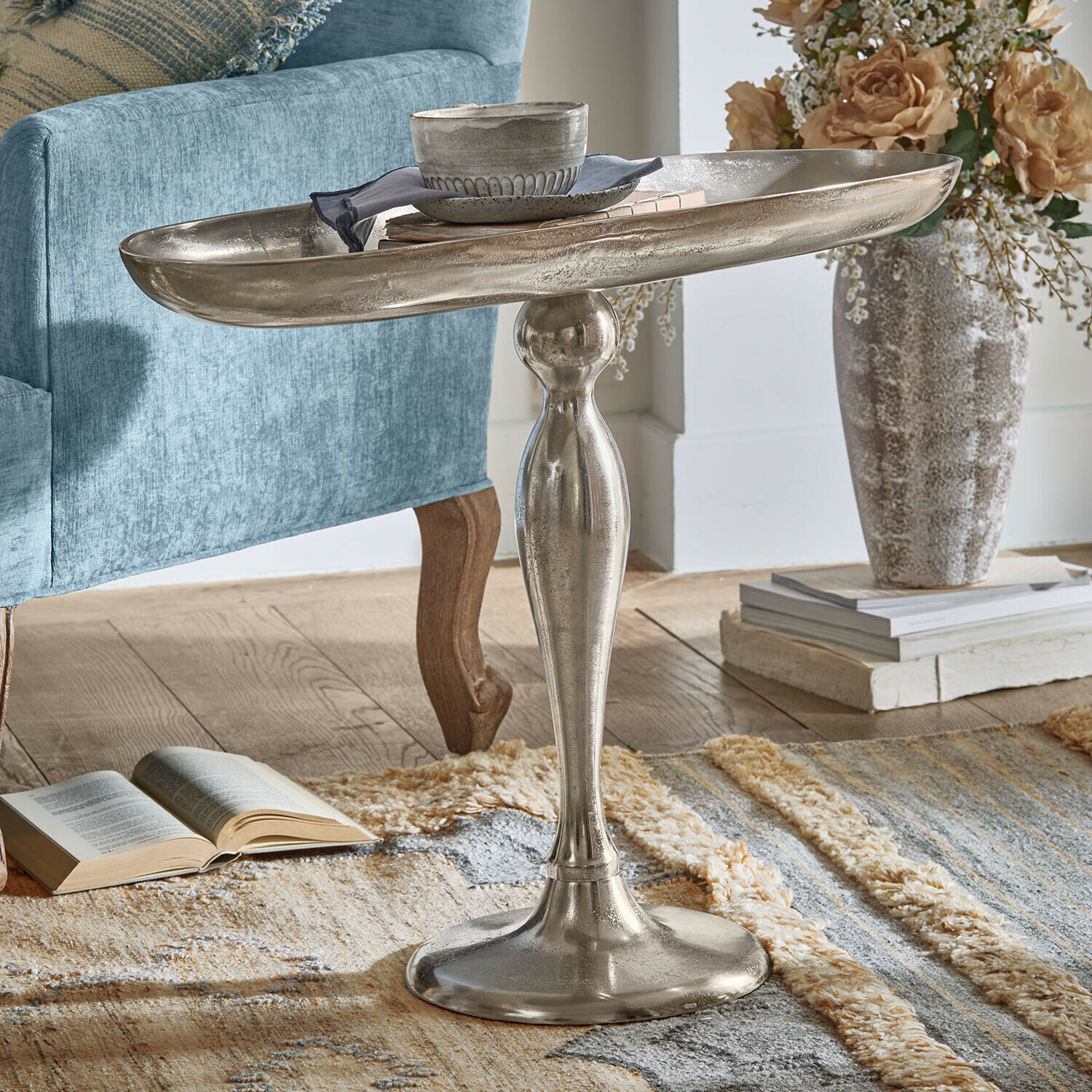 Beistelltisch Stonebrook Silberner Metall-Beistelltisch mit dekorativem Fuß auf Teppich neben blauem Sessel