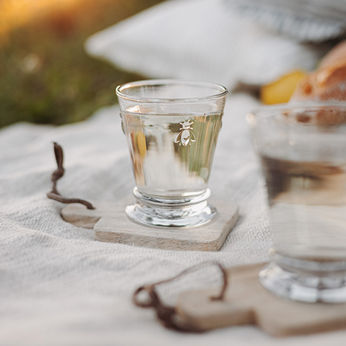 Klares Glas mit Bienenmotiv auf Holzuntersetzer auf Picknickdecke im Freien