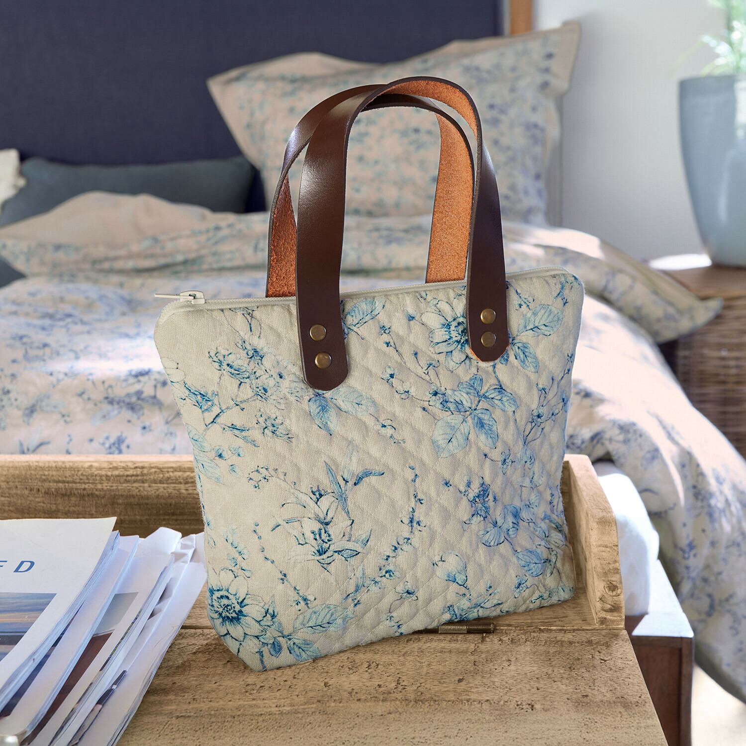 Beige gesteppte Tasche mit blauem Blumenmuster und braunen Ledergriffen auf Holztisch