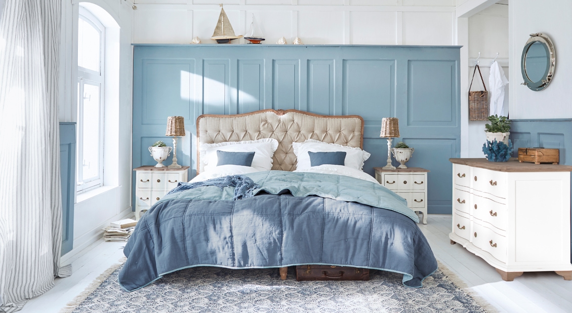 Romantisches Schlafzimmer mit blauer Wand, beige gepolstertem Bett und weißen Holzmöbeln in hellem Ambiente.