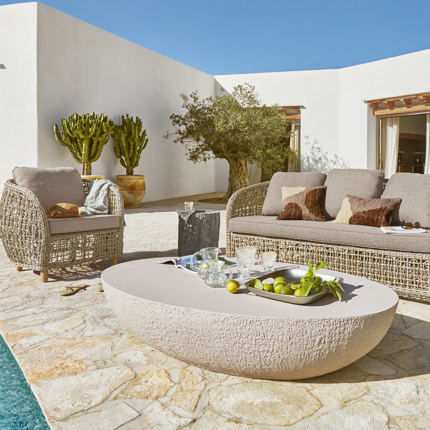 Ovaler Couchtisch aus hellem Stein auf Terrasse mit beigen Rattanmöbeln und grünen Pflanzen