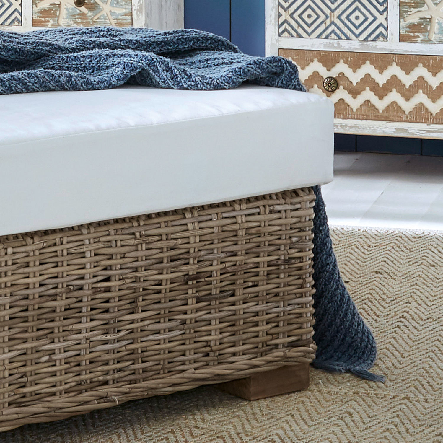 Sitzbank mit weißem Polster auf geflochtener Rattanbasis und blauem Strickdecke auf Juteteppich
