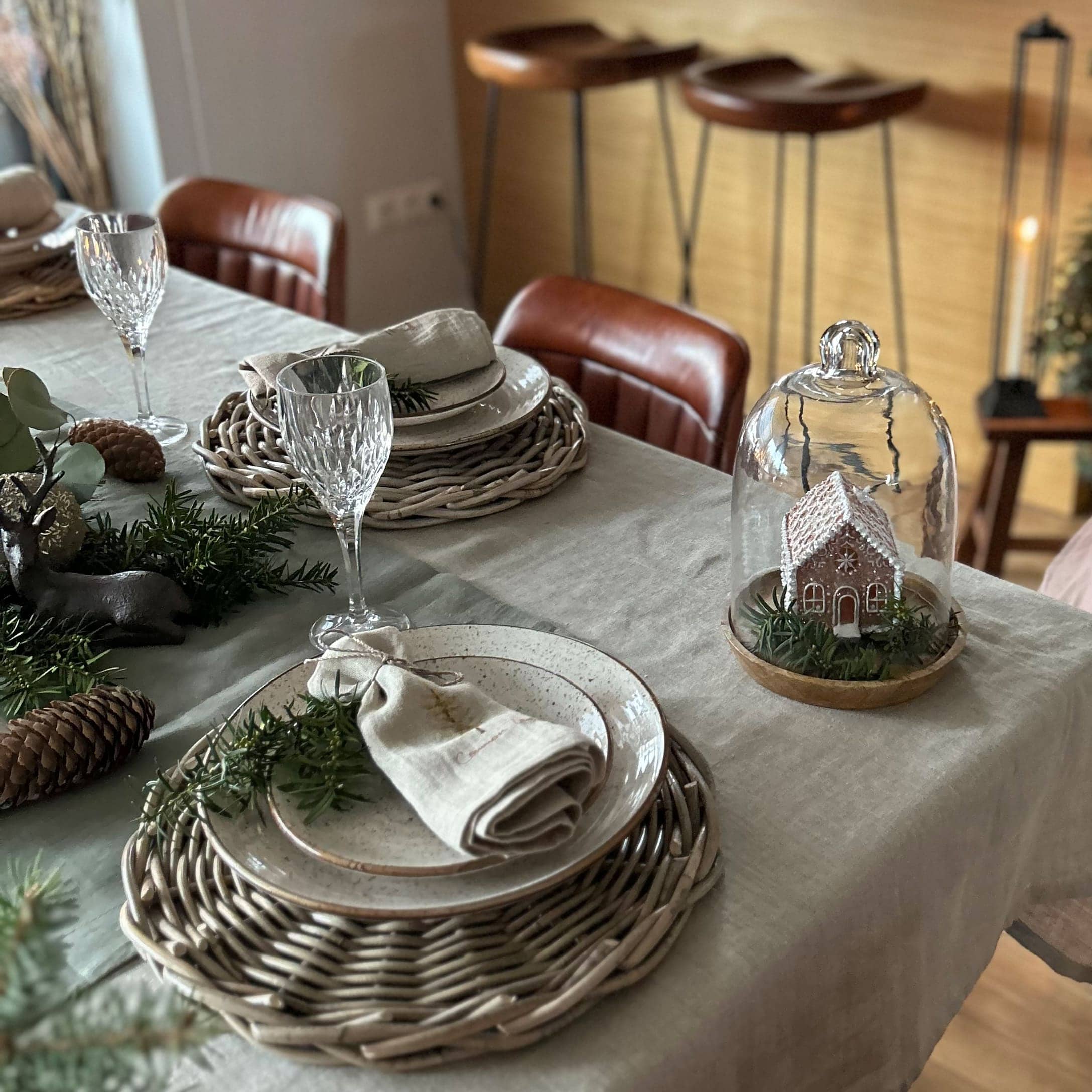 Eleganter gedeckter Tisch mit geflochtenen Platzsets, Keramiktellern und Weihnachtsdeko mit Häuschen unter Glaskuppel