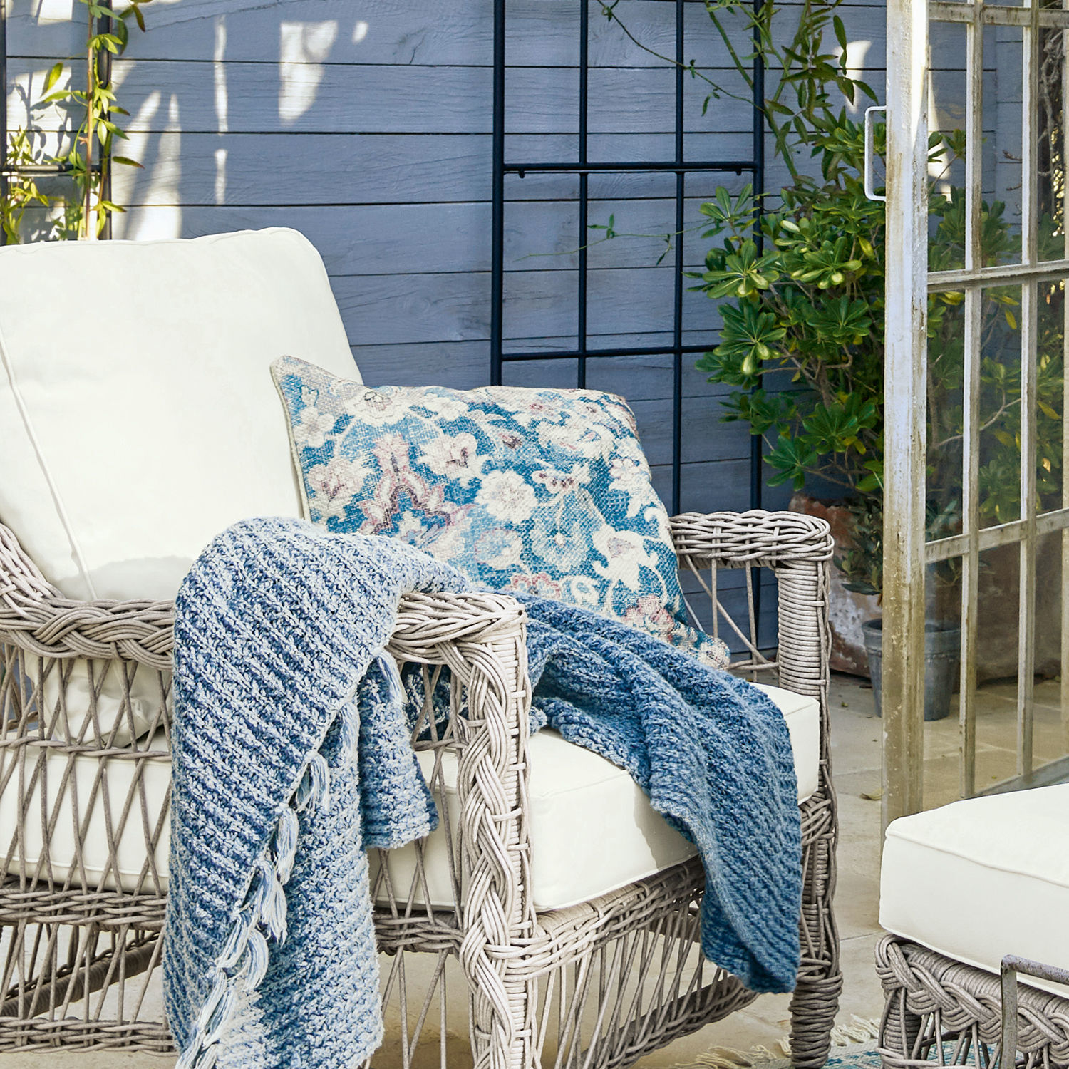 Rattansessel mit cremefarbenen Kissen und blauem Strickplaid in gemütlichem Garten