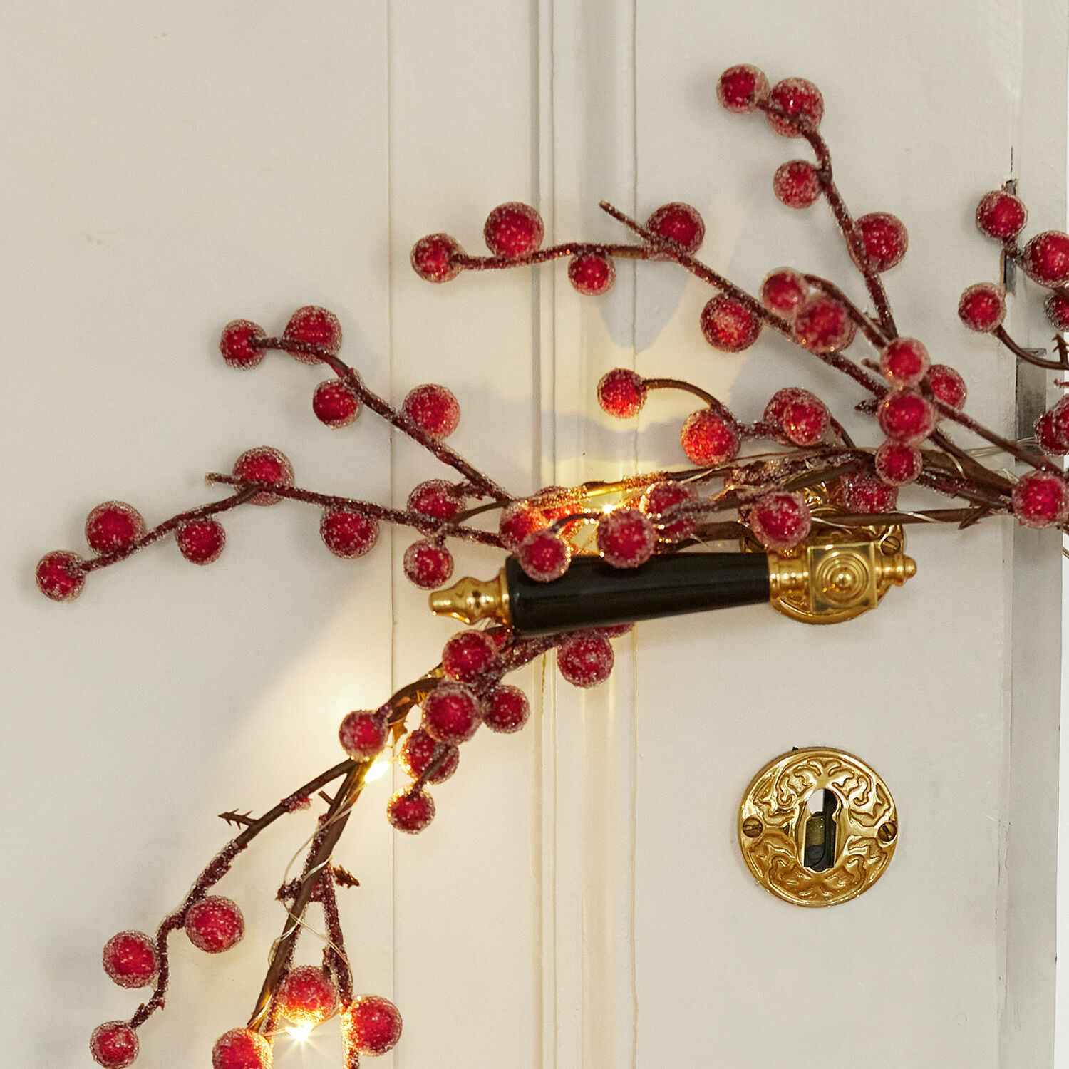 Zweig mit roten Beeren und Lichterkette an weißer Tür mit schwarz-goldenem Türgriff