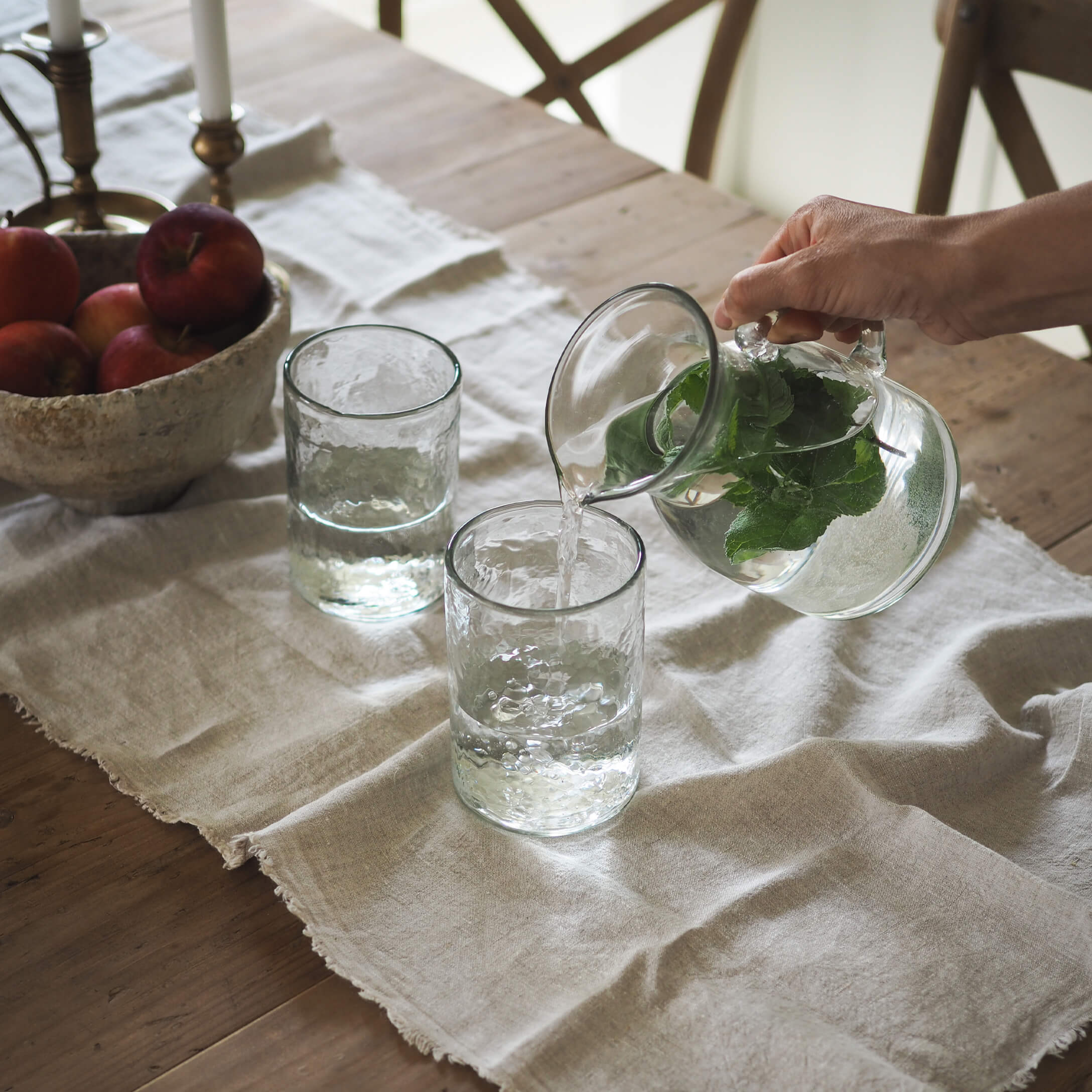 Glaskrug mit grünen Blättern und zwei Gläser auf einem Leinentuch auf einem Holztisch