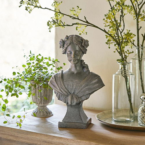 Graue Steinskulptur einer Frau mit Blumen im Haar auf Holztisch mit Pflanzenarrangement