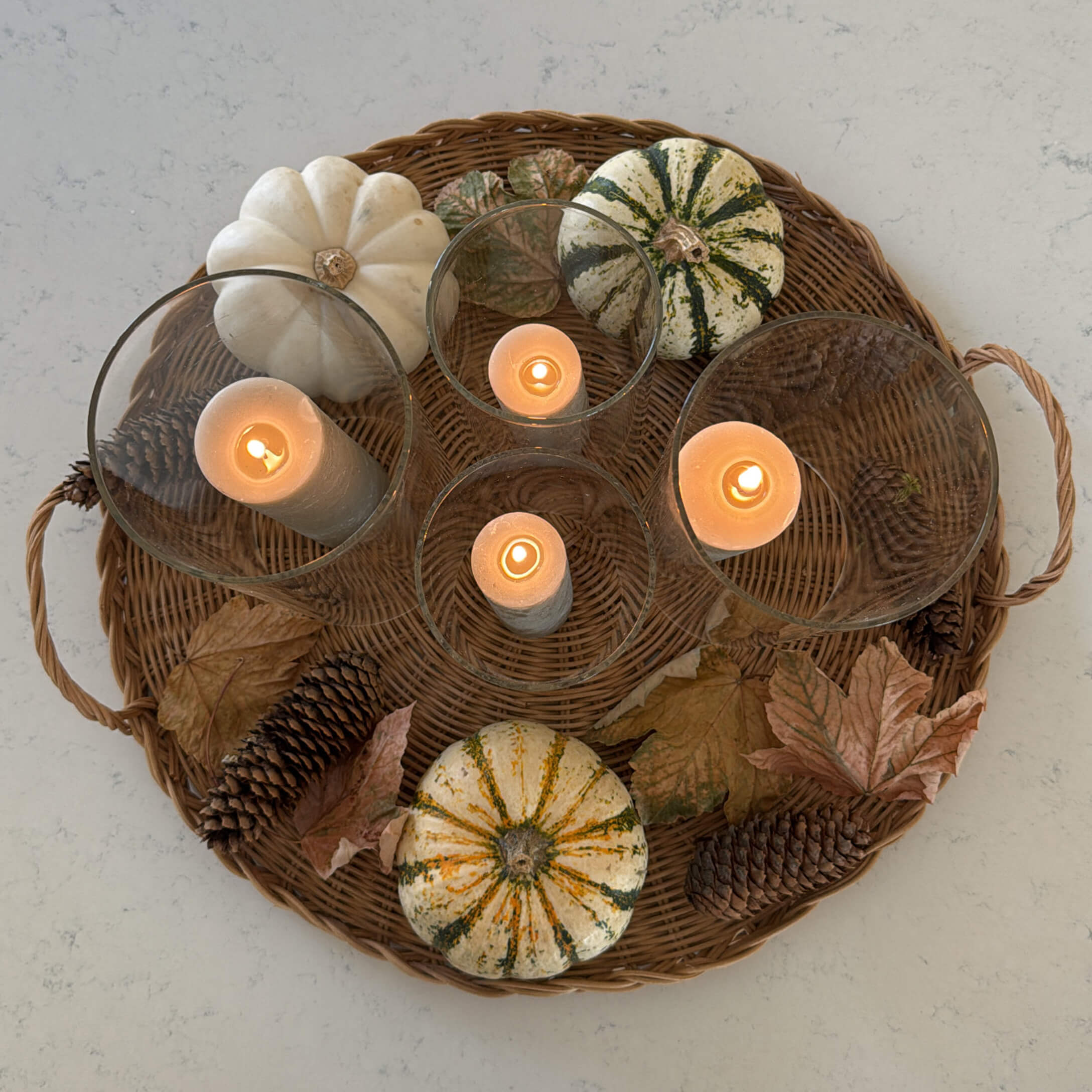 Herbstliches Ensemble mit Kürbissen, Tannenzapfen und brennenden Kerzen in Glas auf einem geflochtenen Tablett