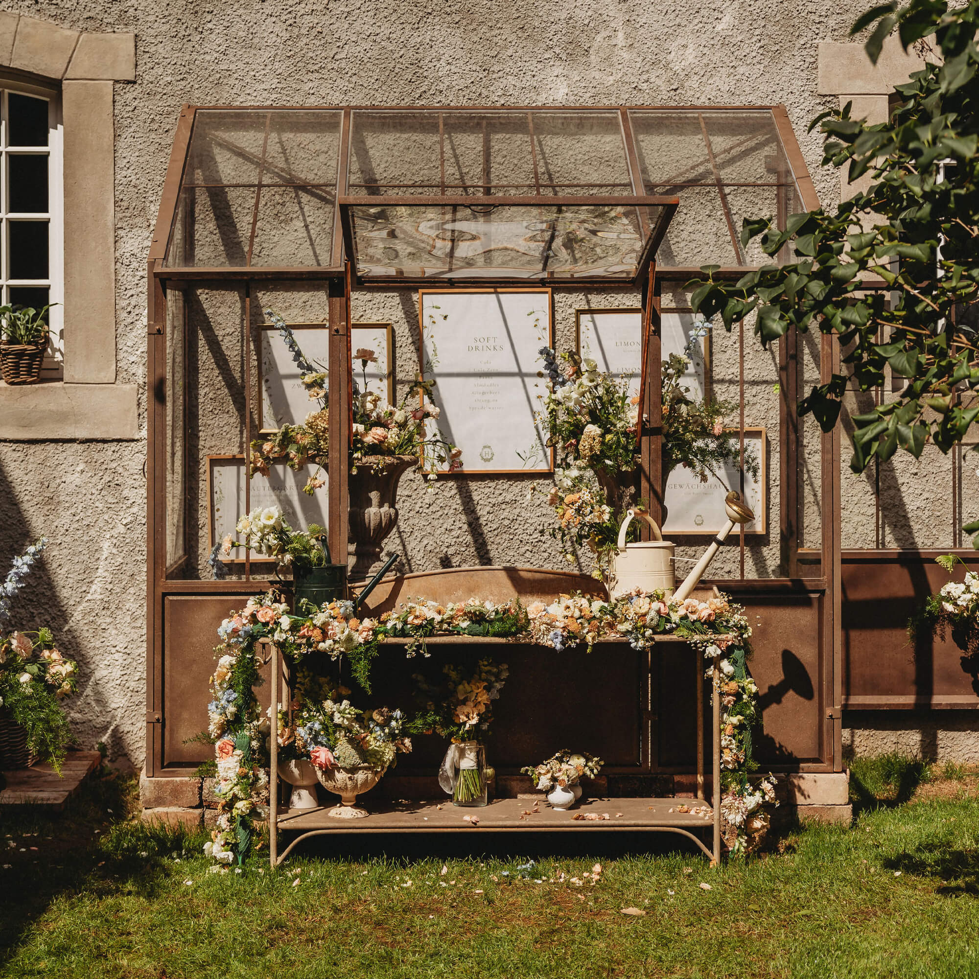 Metall-Gartentisch mit Blumenarrangements und Gießkannen vor einer Glashauswand
