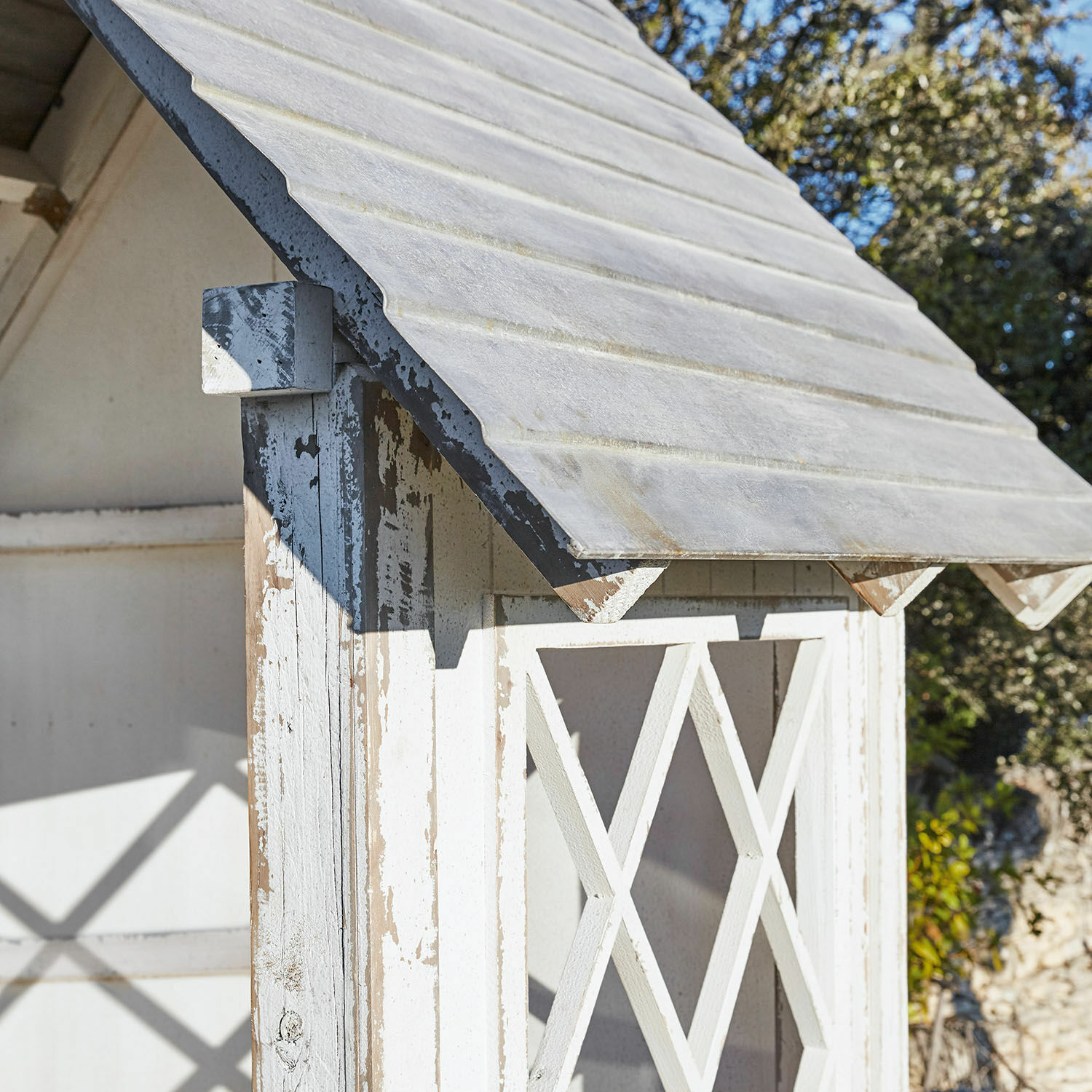 Weißes Holz-Gartenhaus mit abgenutztem Look und grauem Dach vor blauem Himmel und Bäumen