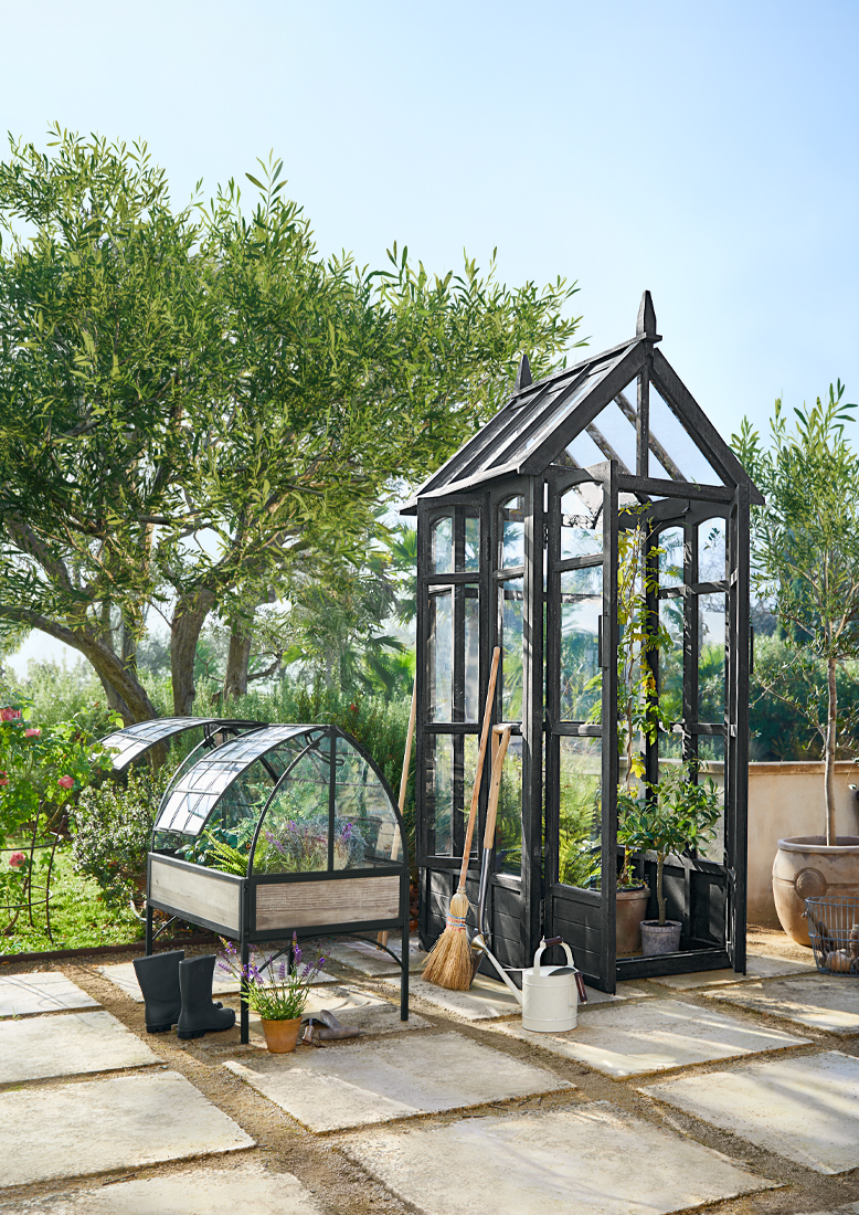 Schwarzes Gartenhaus aus Metall und Glas mit Gartengeräten auf sonniger Terrasse mit Pflanzen.