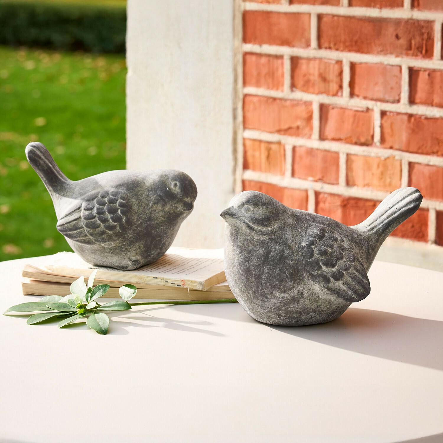Zwei graue Vogel-Figuren aus Stein auf weißem Tisch mit Buch und grünem Blatt als Dekoration
