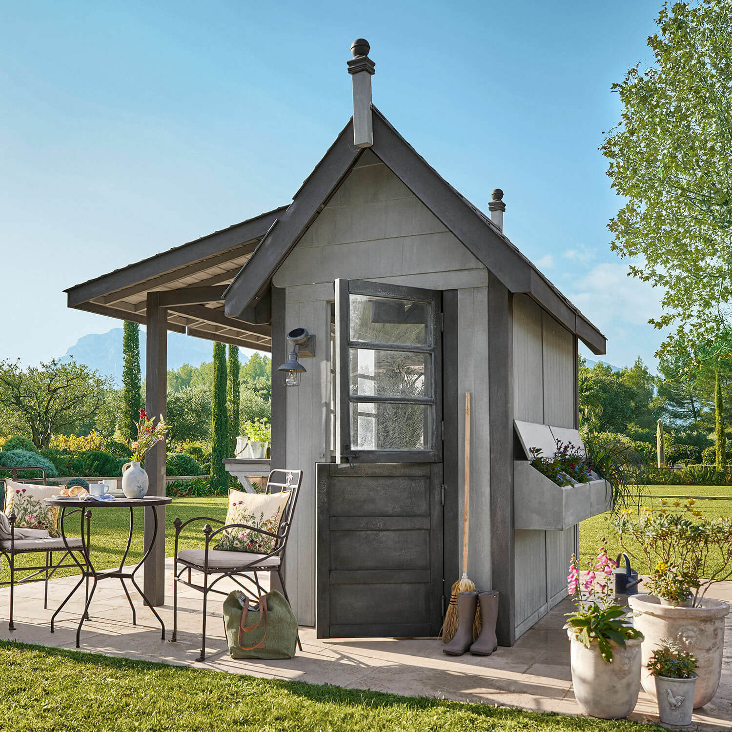 Graue Holz-Gartenlaube mit schwarzer Tür, Blumenkästen und Metall-Gartenmöbeln im Grünen