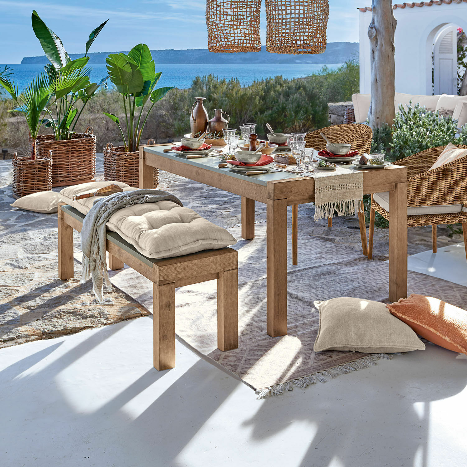 Holztisch und Bank mit beigen Kissen auf Terrasse mit Meerblick und Pflanzen in Korbgefäßen