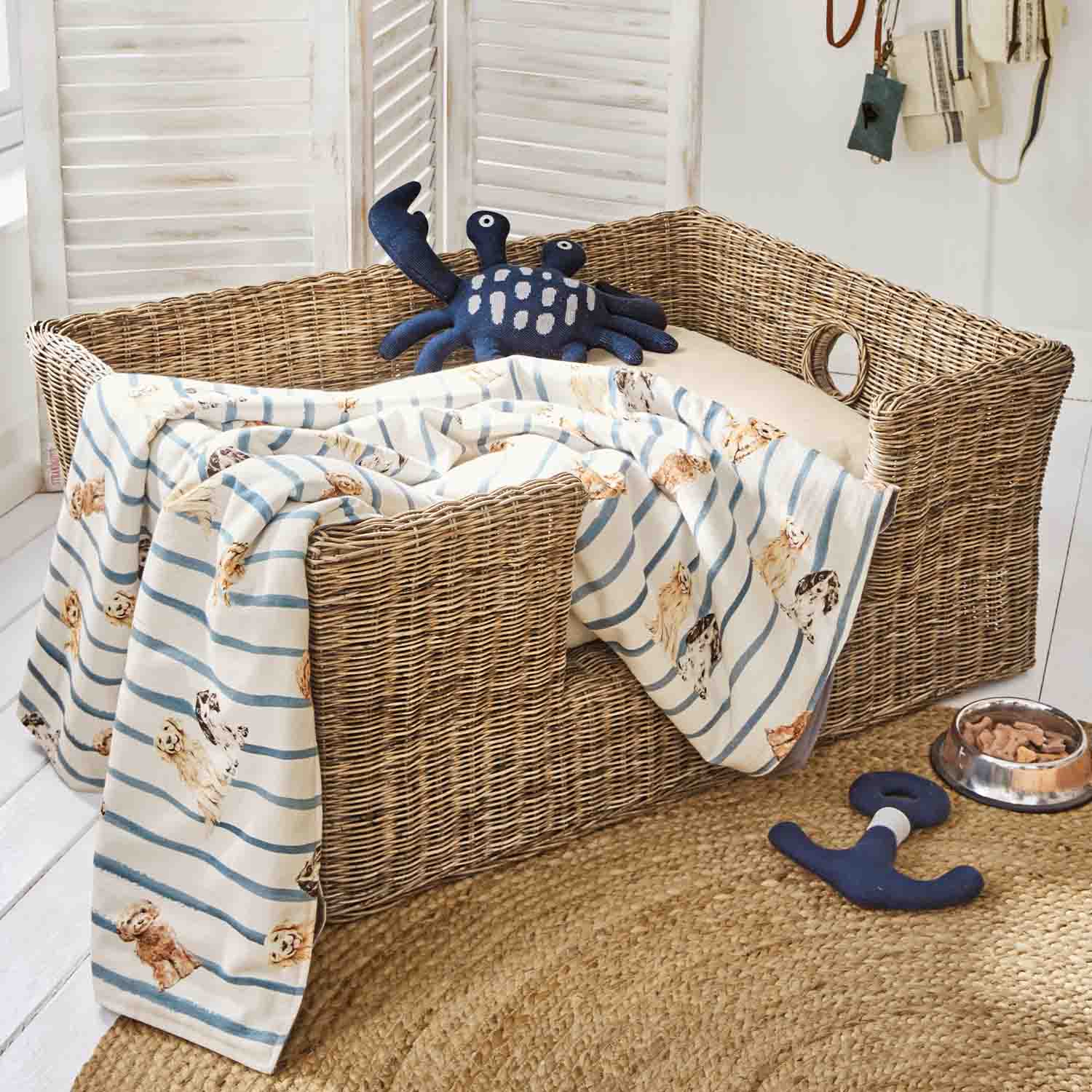 Weiden-Hundebett mit cremefarbenem Kissen, gestreifter Decke mit Hundemotiven und dunkelblauem Krabben-Spielzeug
