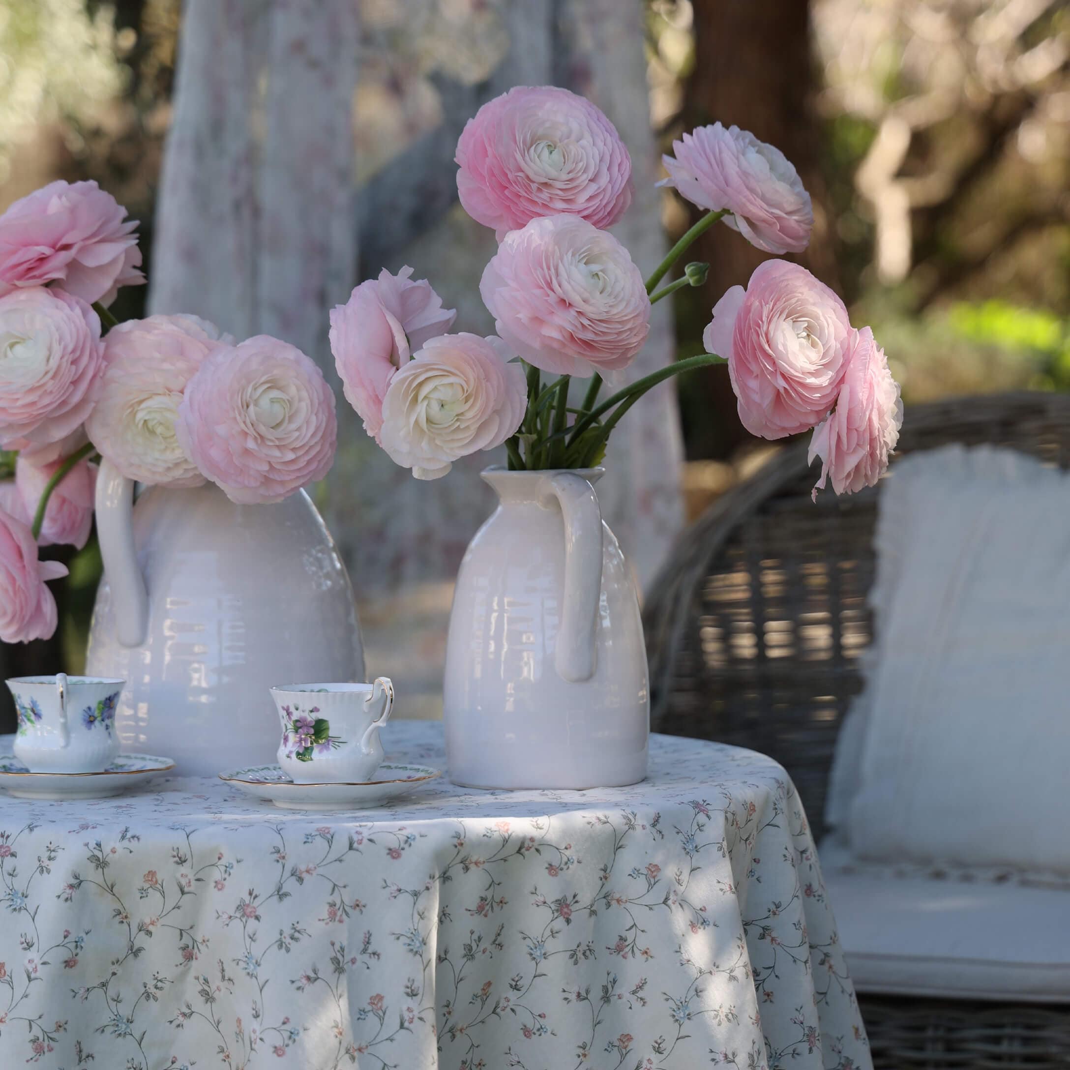 Weiße Keramikkrüge mit rosa Blumen auf einem Tisch mit geblümtem Tischtuch und dekorativen Teetassen