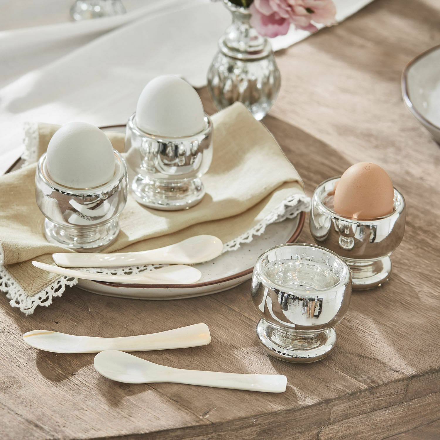 Eierbecher 4er Set Lilligret Set aus silbernen Metall-Eierbechern mit weißen und braunen Eiern und weißen Löffeln auf Holztisch