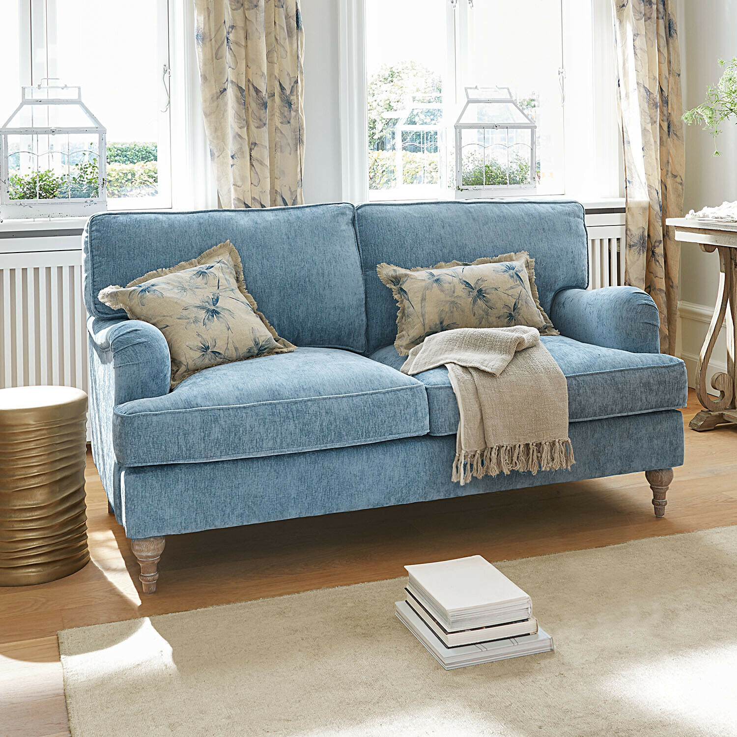 Sofa Elvika Blaues Stoffsofa mit floralen Kissen und beiger Decke in hellem Wohnzimmer