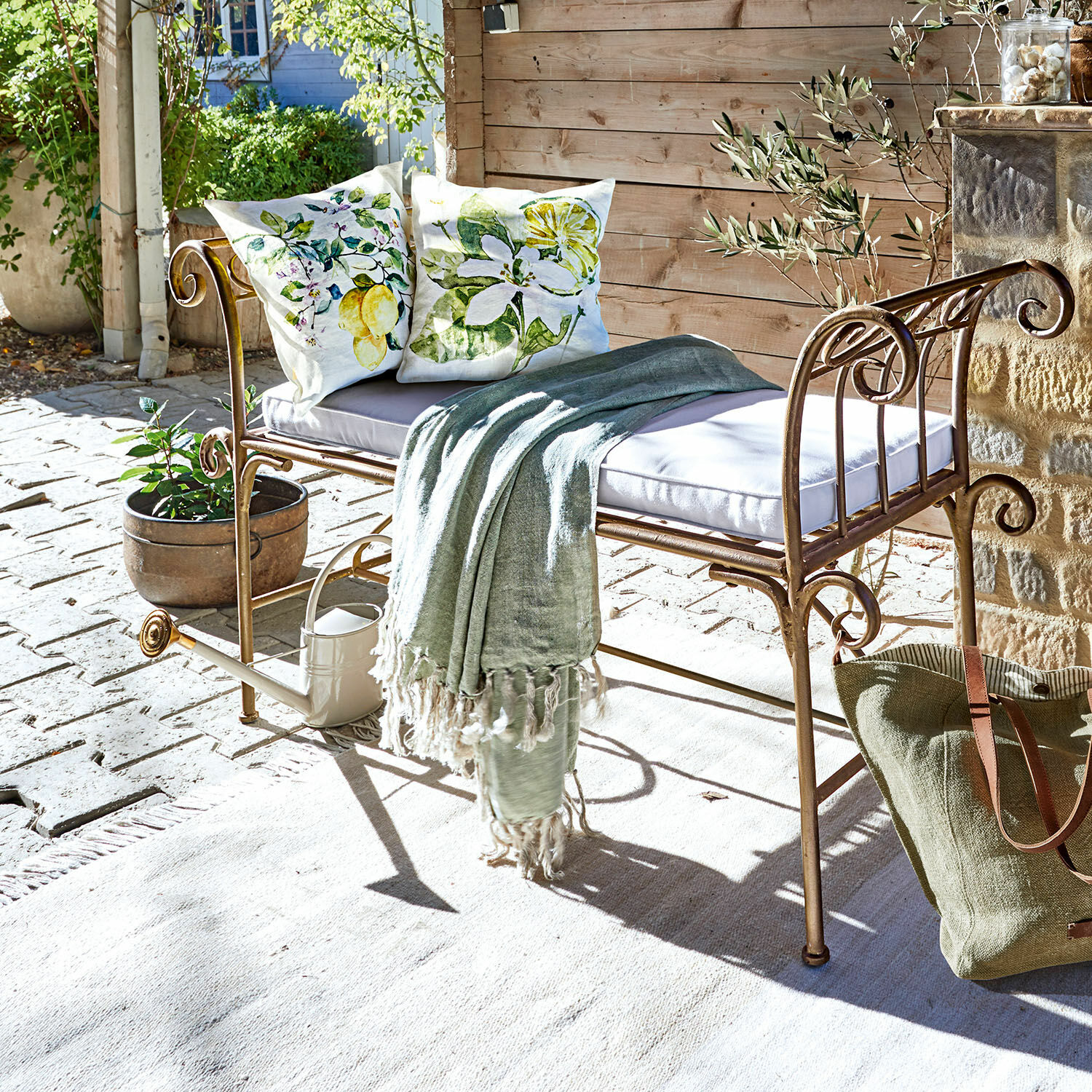 Metall-Gartenbank mit Polster und grünem Plaid auf hellem Teppich auf sonniger Terrasse