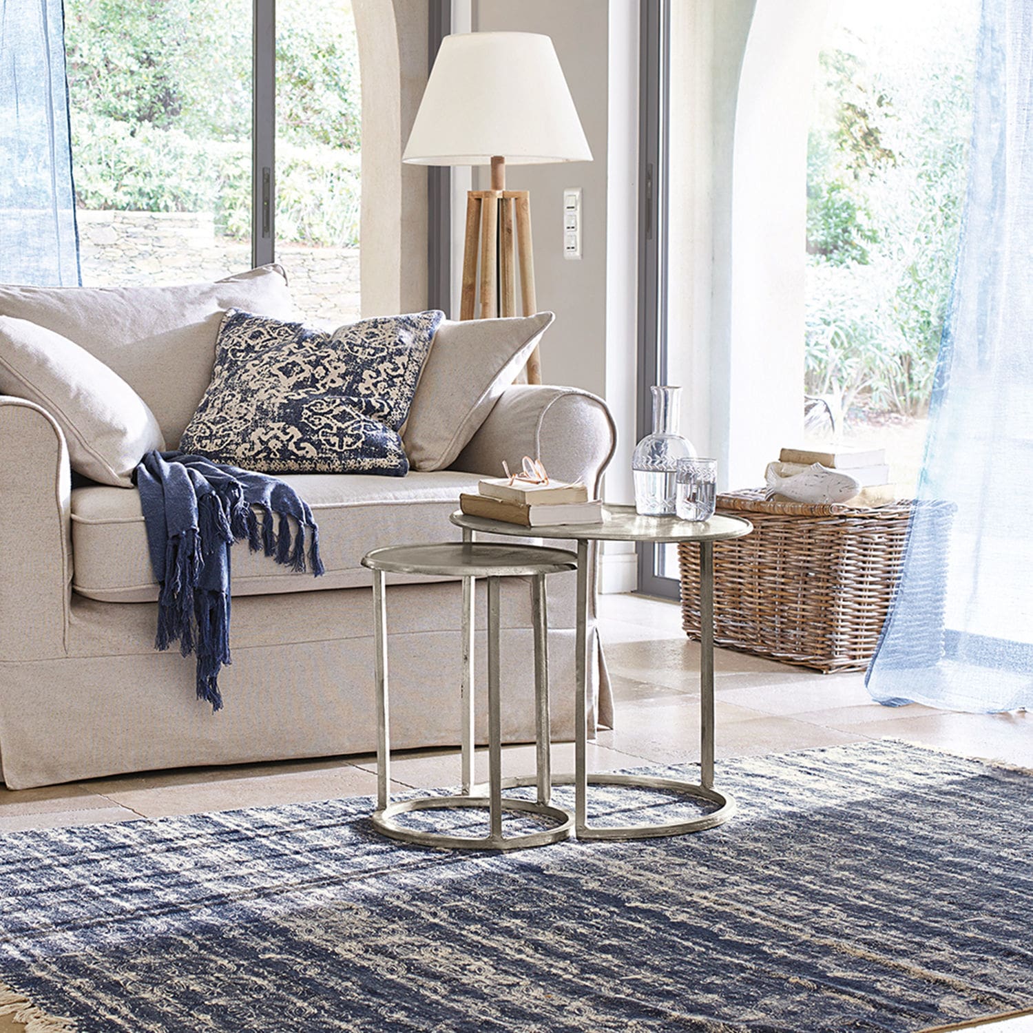 Beiges Sofa mit blauem Kissen und Decke, zwei Metall-Couchtische auf blauem Teppich in hellem Wohnzimmer