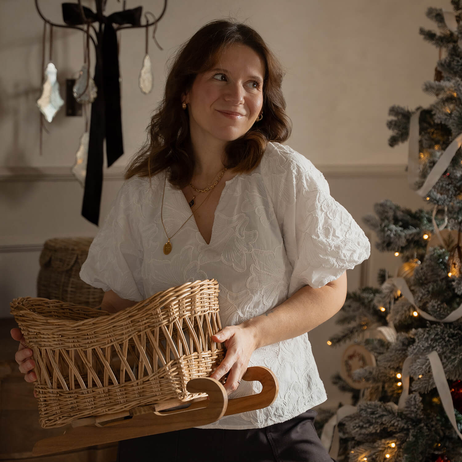 Frau in weißer Bluse hält einen Korbschlitten aus Weide mit Holzgestell neben geschmücktem Weihnachtsbaum
