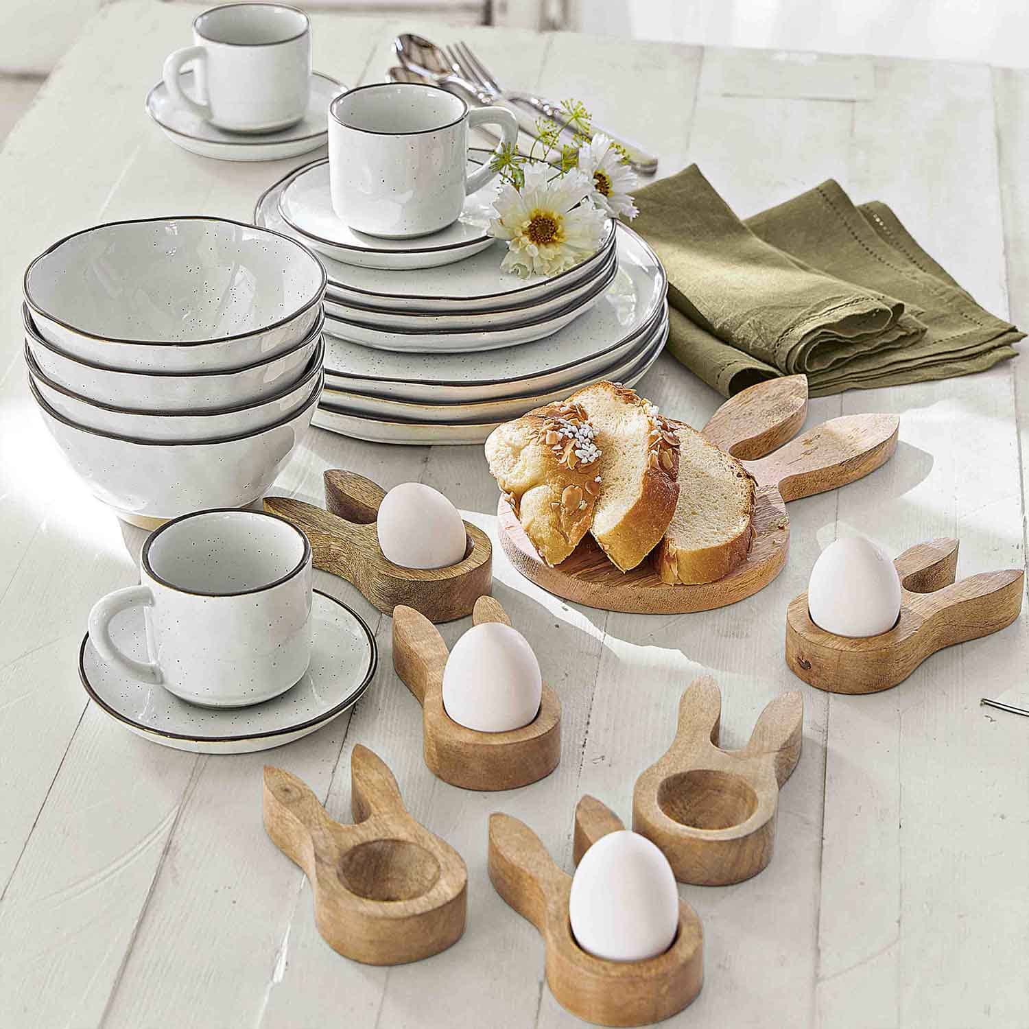 Weißes Keramikgeschirr mit braunem Rand und hölzernen Eierhaltern in Hasenform für den Frühstückstisch