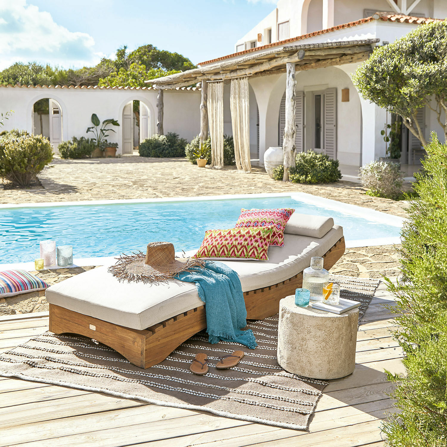 Gartenliege aus Holz mit cremefarbenem Polster und bunten Kissen am Pool auf der Terrasse