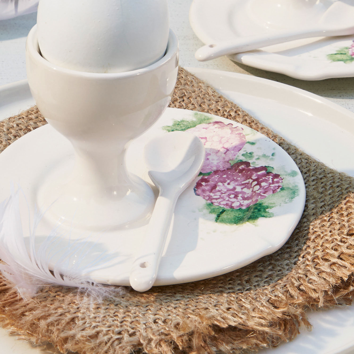 Weißer Keramik-Eierbecher mit Löffel und Teller mit Blumenmuster auf Juteunterlage