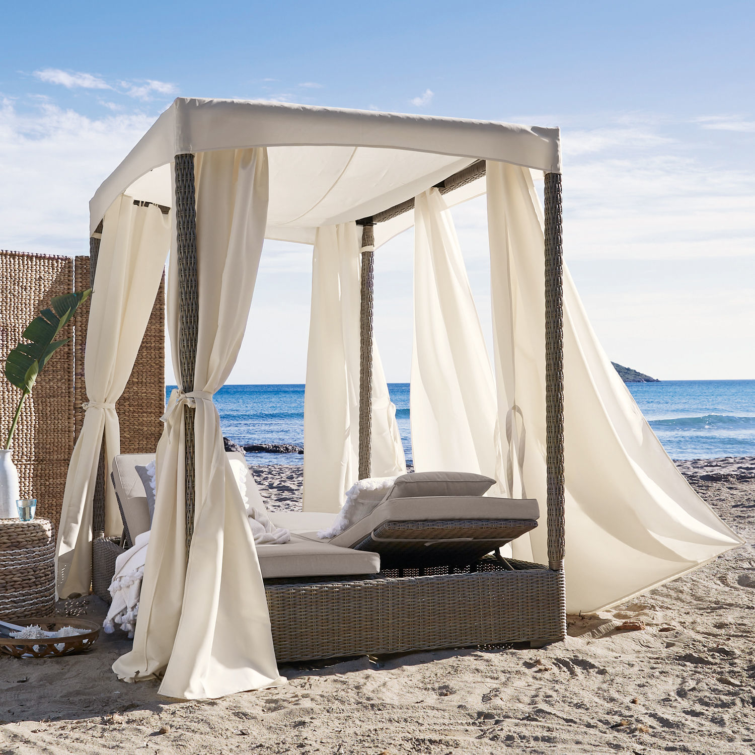 Rattan-Liegestuhl mit cremefarbenen Vorhängen und Kissen am Strand mit Meerblick