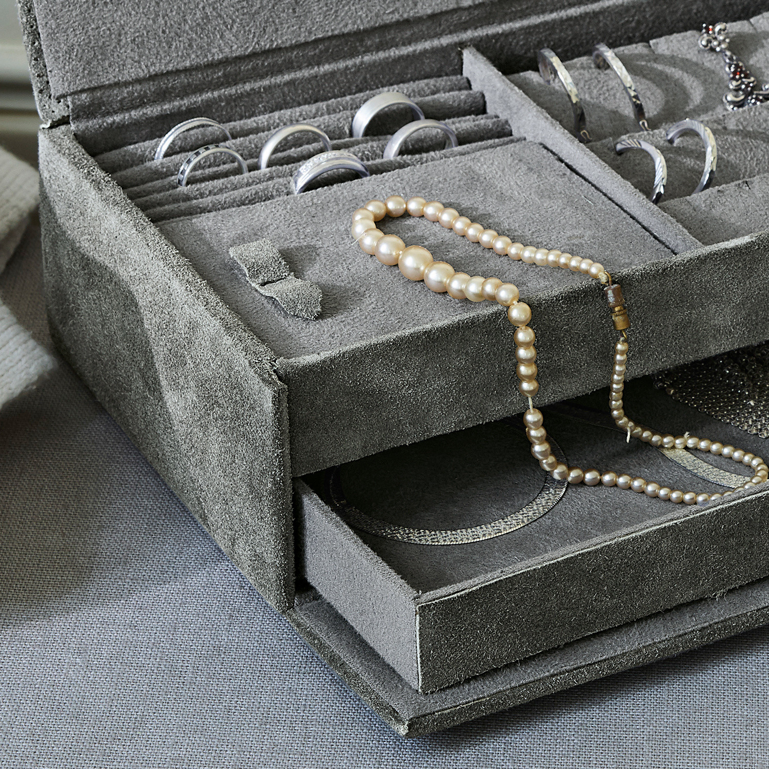 Grauer Schmuckkasten aus Wildleder mit Perlenkette und silbernen Ringen ordentlich verstaut