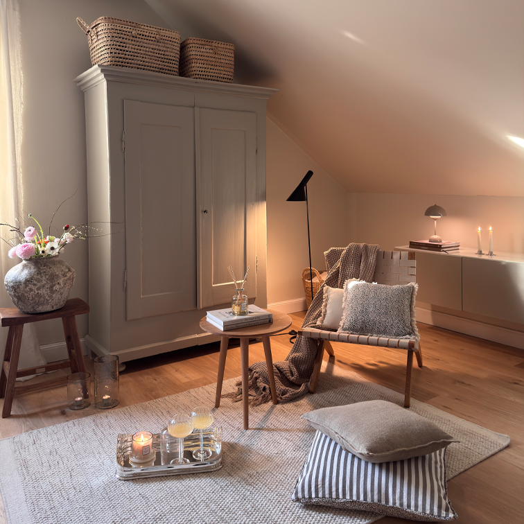 Gemütliches Wohnzimmer mit beigem Sessel, Holztisch und Schrank unter Dachschräge, Dekorationen und Lampen