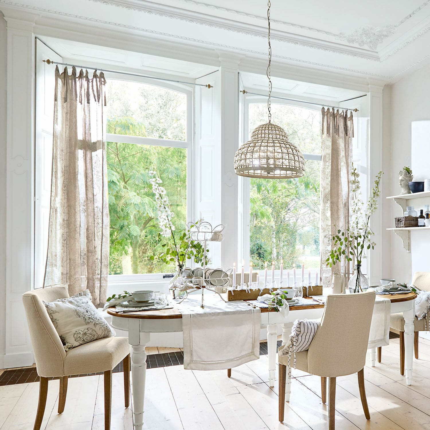 Esszimmer mit weißem Holztisch, beigen Stühlen, naturfarbenen Vorhängen und Blumenarrangements