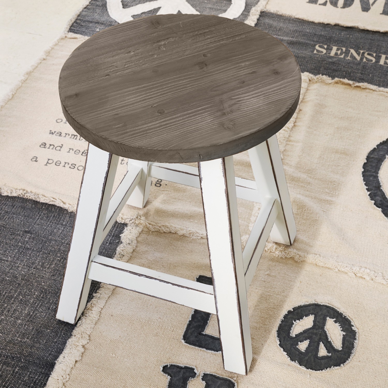 Holzhocker mit rundem braunem Sitz und weißen Beinen auf gemustertem Teppich mit Friedenssymbol.
