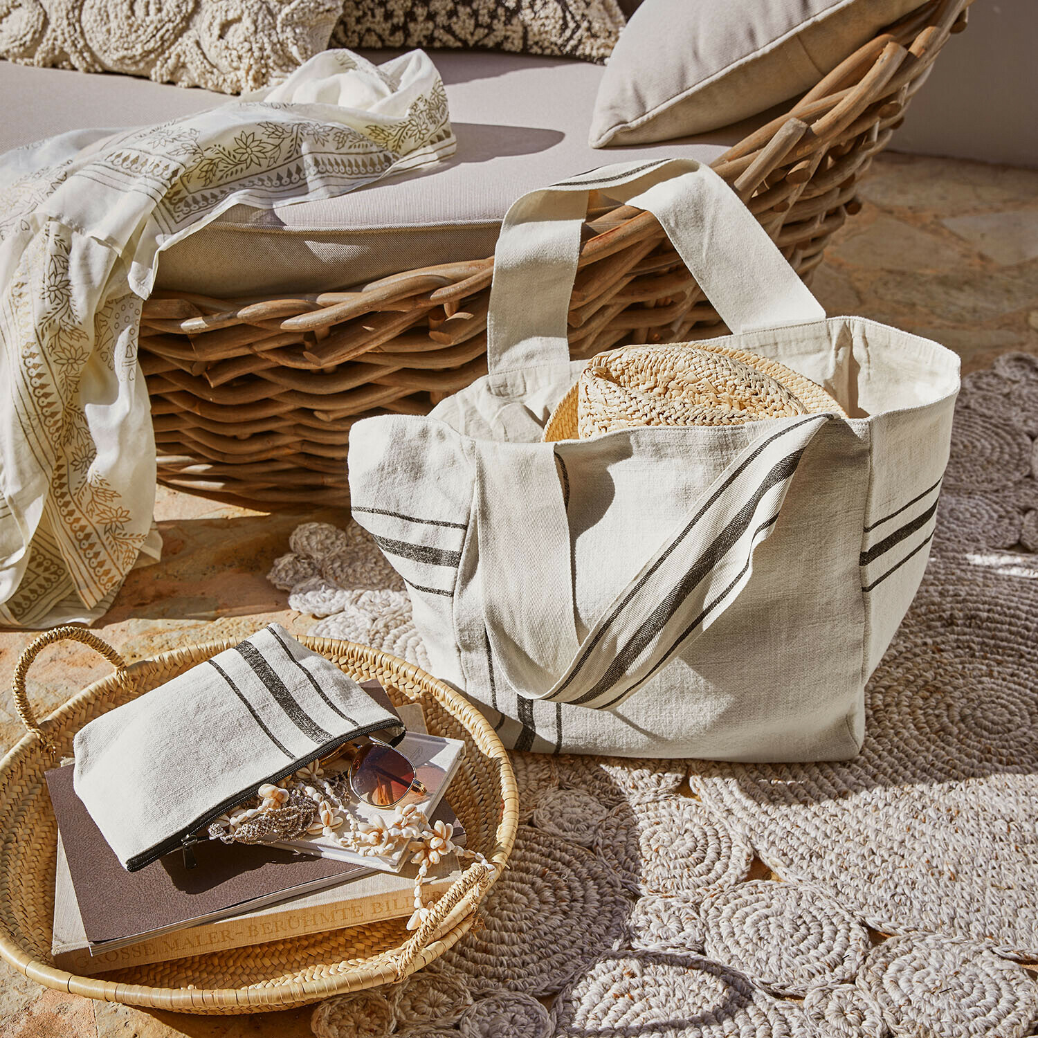 Beige Baumwolltasche mit Streifen und Strohhut vor Korbsessel und Teppich im Sonnenlicht