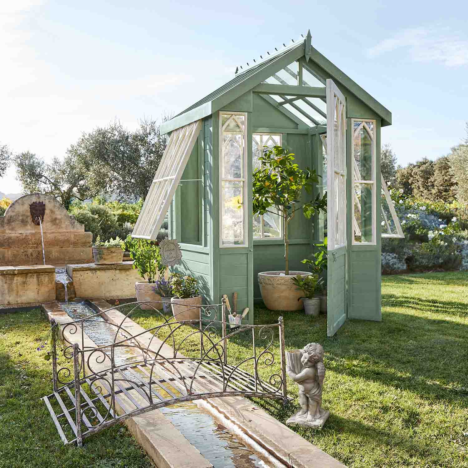 Grünes Holz-Gewächshaus mit weißen Fenstern im Garten mit Pflanzen und Brunnen