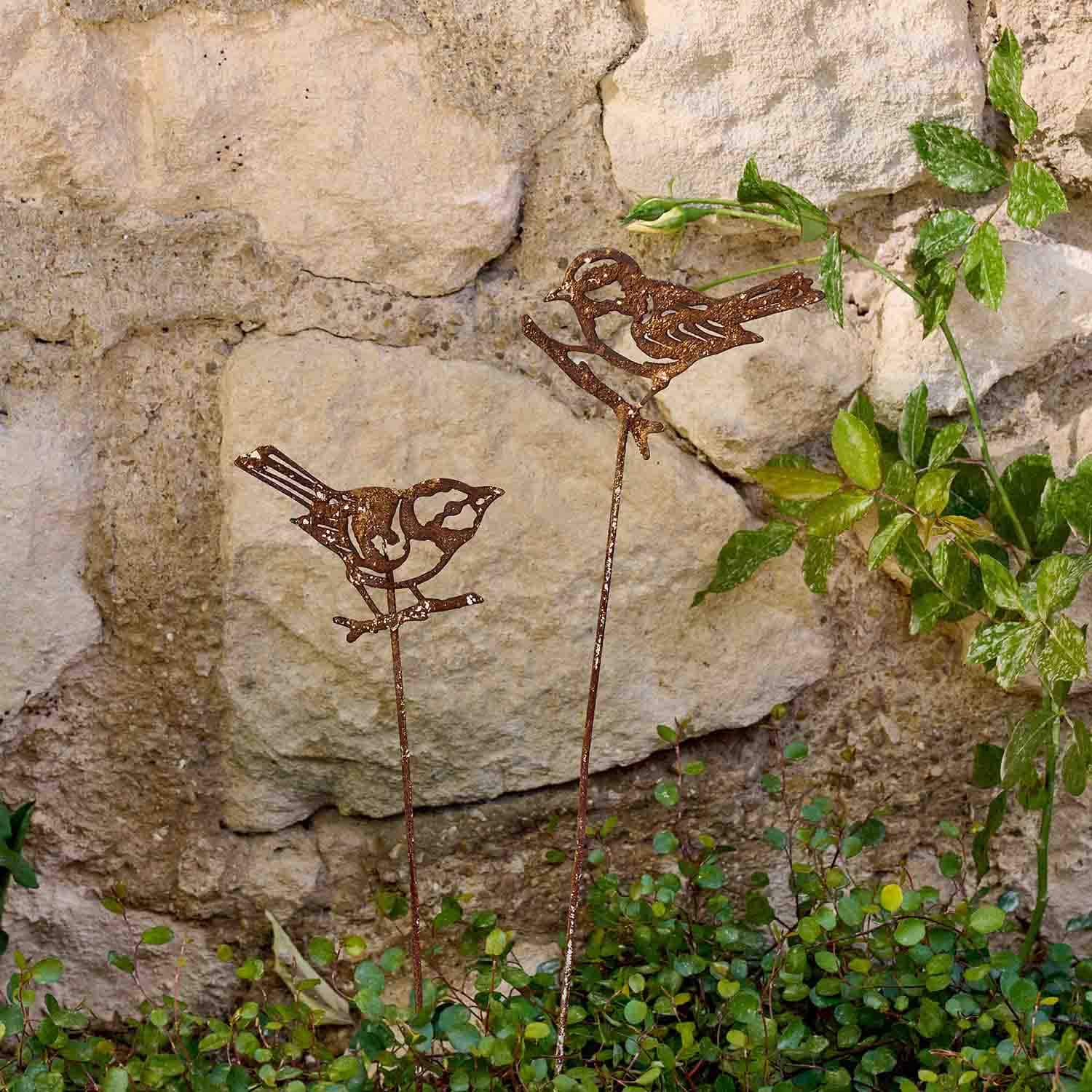 Dekorative Metallvögel in Rostoptik auf Stäben im Garten vor einer Steinmauer