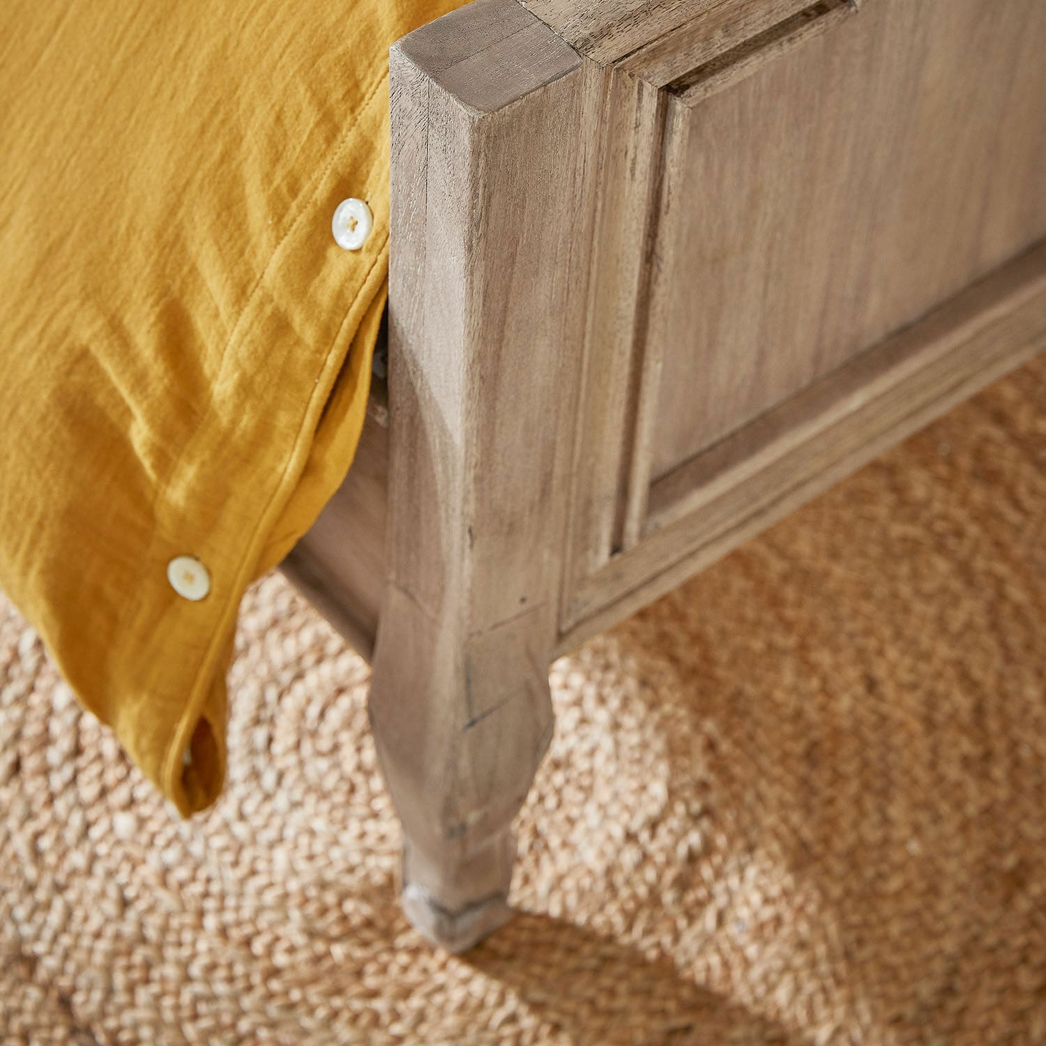Eckdetail eines naturbelassenen Holzbetts mit gelber Baumwollbettdecke auf Jute-Teppich