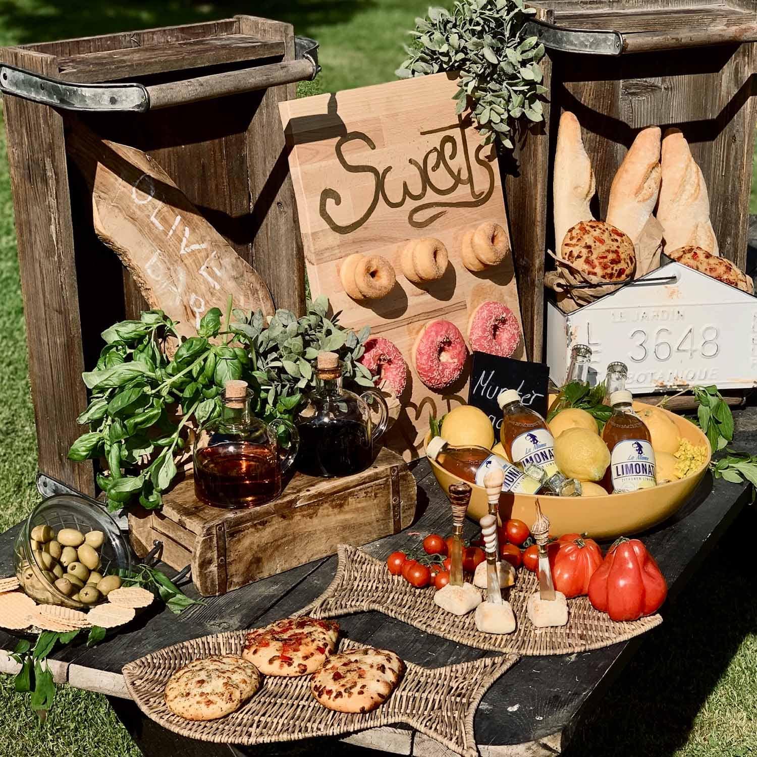 Rustikaler Tisch mit Holzkisten, Brot, Oliven, Kräutern und Limonade im Freien bei Sonnenschein