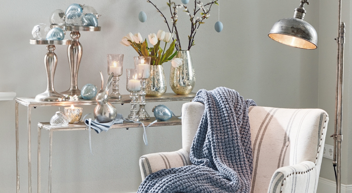 Gemütlicher weißer Sessel mit blauem Strickplaid und silbernen Kerzenhaltern sowie Dekorationen im Hintergrund