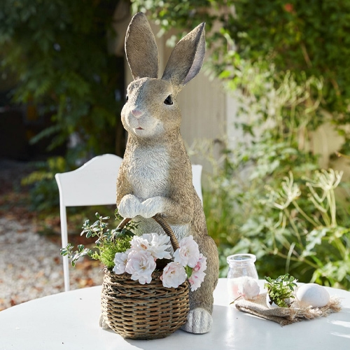 Dekorativer Hase aus Kunststoff in Naturfarben mit Weidenkorb und Blumen auf Gartentisch