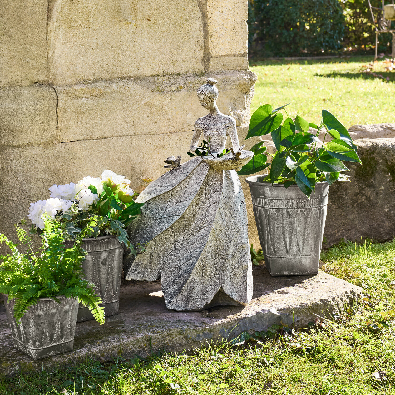 Gartendekoration aus Stein in Form einer Frau mit Blätterkleid und Pflanzgefäßen mit grünen Pflanzen