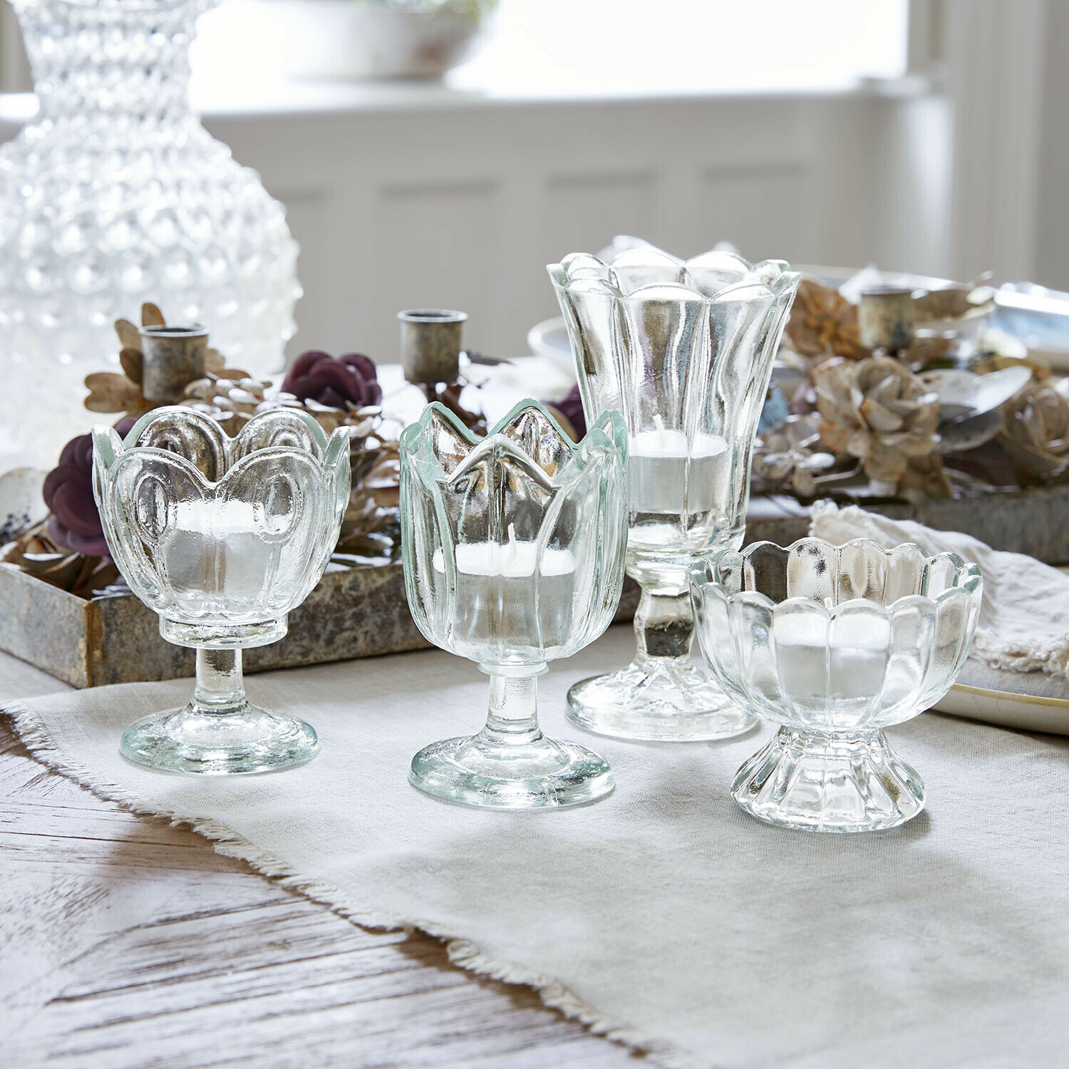 Set aus vier transparenten Glas-Teelichthaltern in verschiedenen Formen auf hellem Tuch