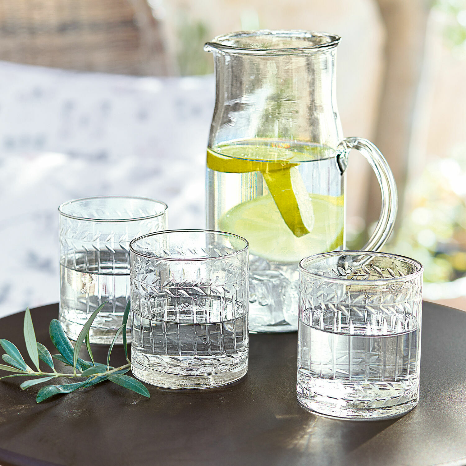 Glas 6er Set Padiès Glaskrug mit Zitronenscheiben und drei Wassergläser auf dunklem Tisch mit grünen Blättern als Dekor