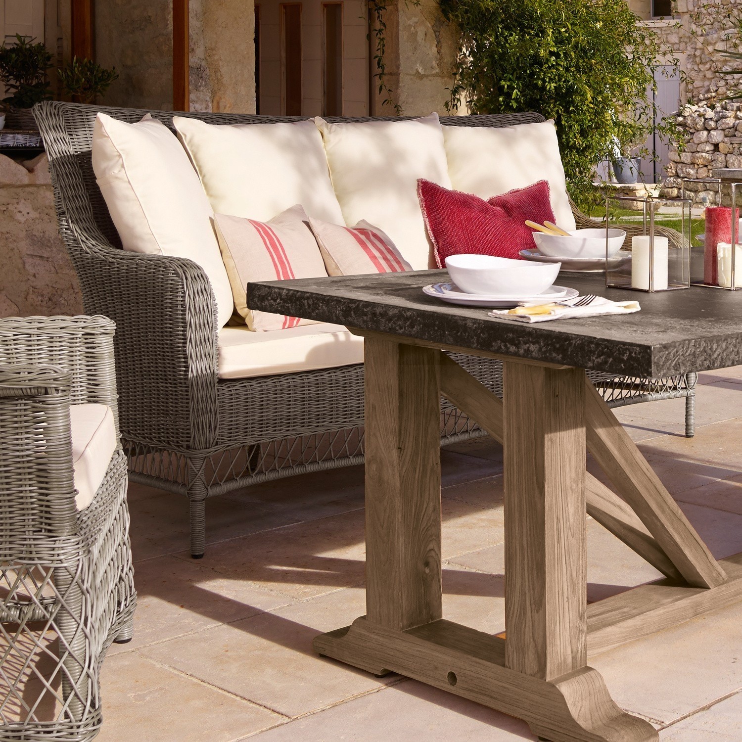 Graues Rattan-Sofa mit cremefarbenen Kissen und Holztisch mit schwarzer Tischplatte auf Terrasse