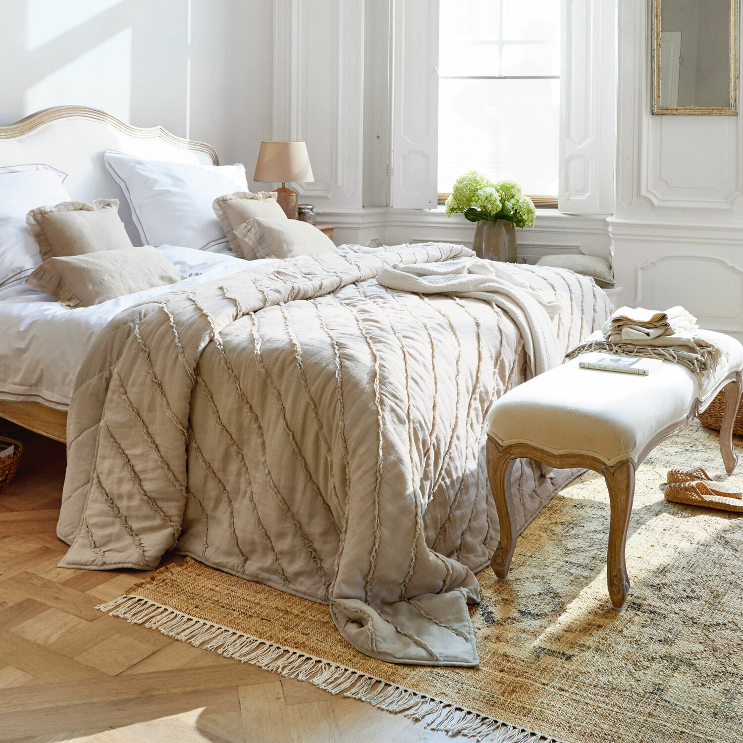 Beige Baumwollbettdecke auf Bett in hellem Schlafzimmer mit dekorativen Kissen und Holzbank