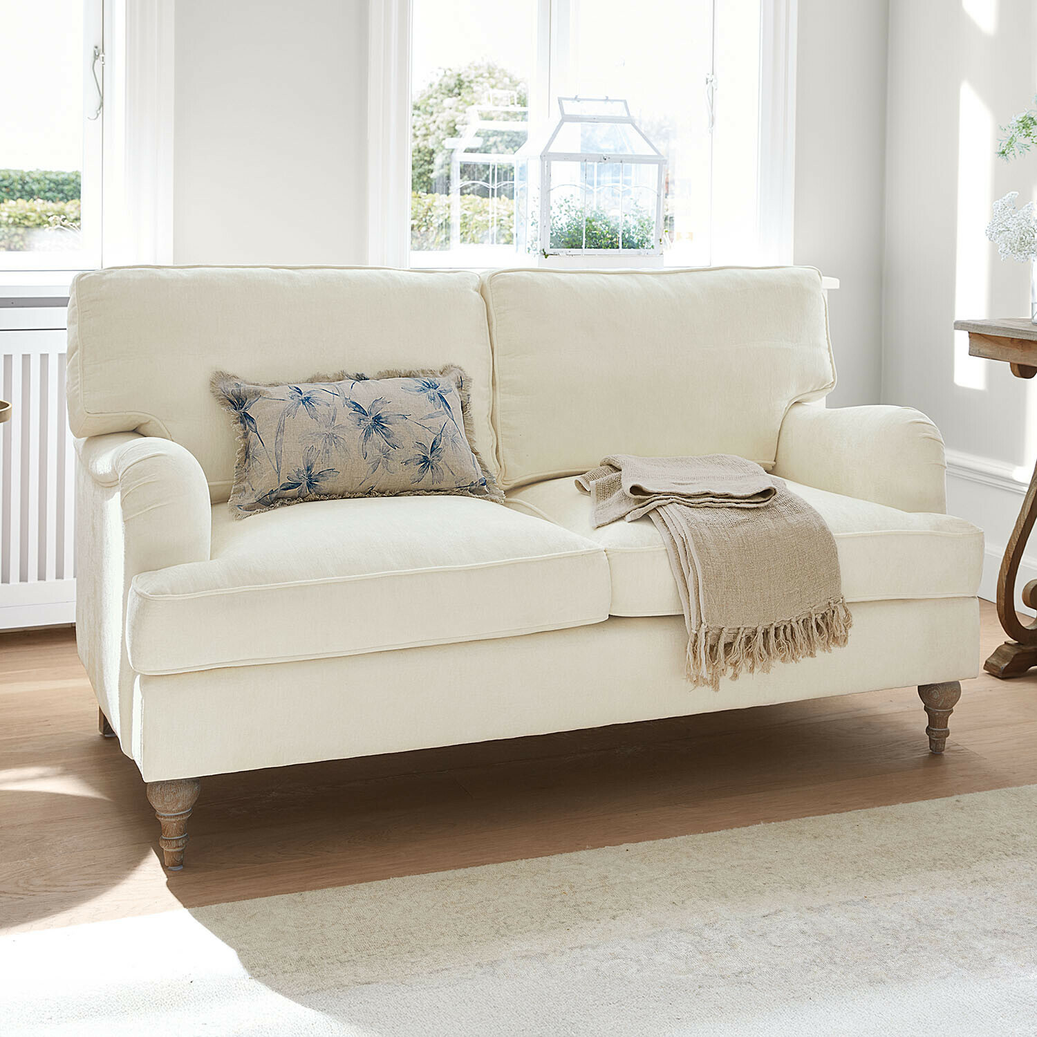 Sofa Elvika Beige bezogene Couch mit floralem Kissen und beigem Plaid in hellem Wohnzimmer