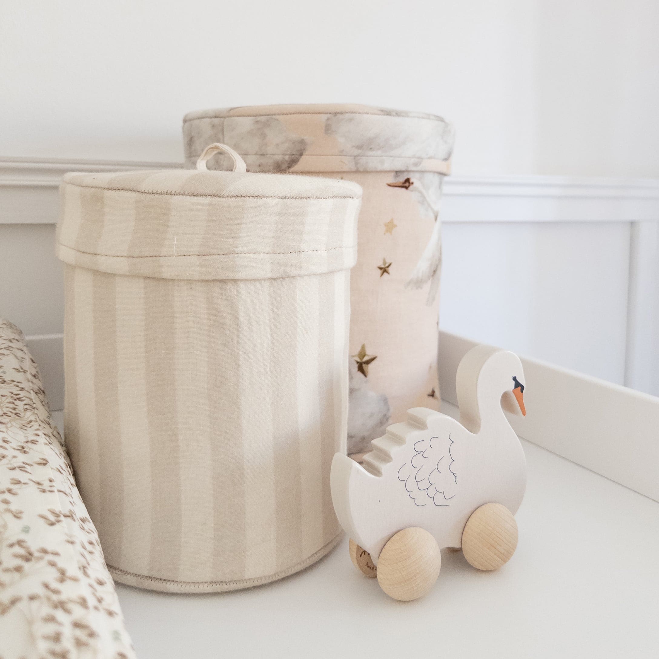 Zwei Stoffbehälter in Beige mit Streifen und Mustern sowie ein hölzernes Schwan-Spielzeug auf Rädern fürs Kinderzimmer
