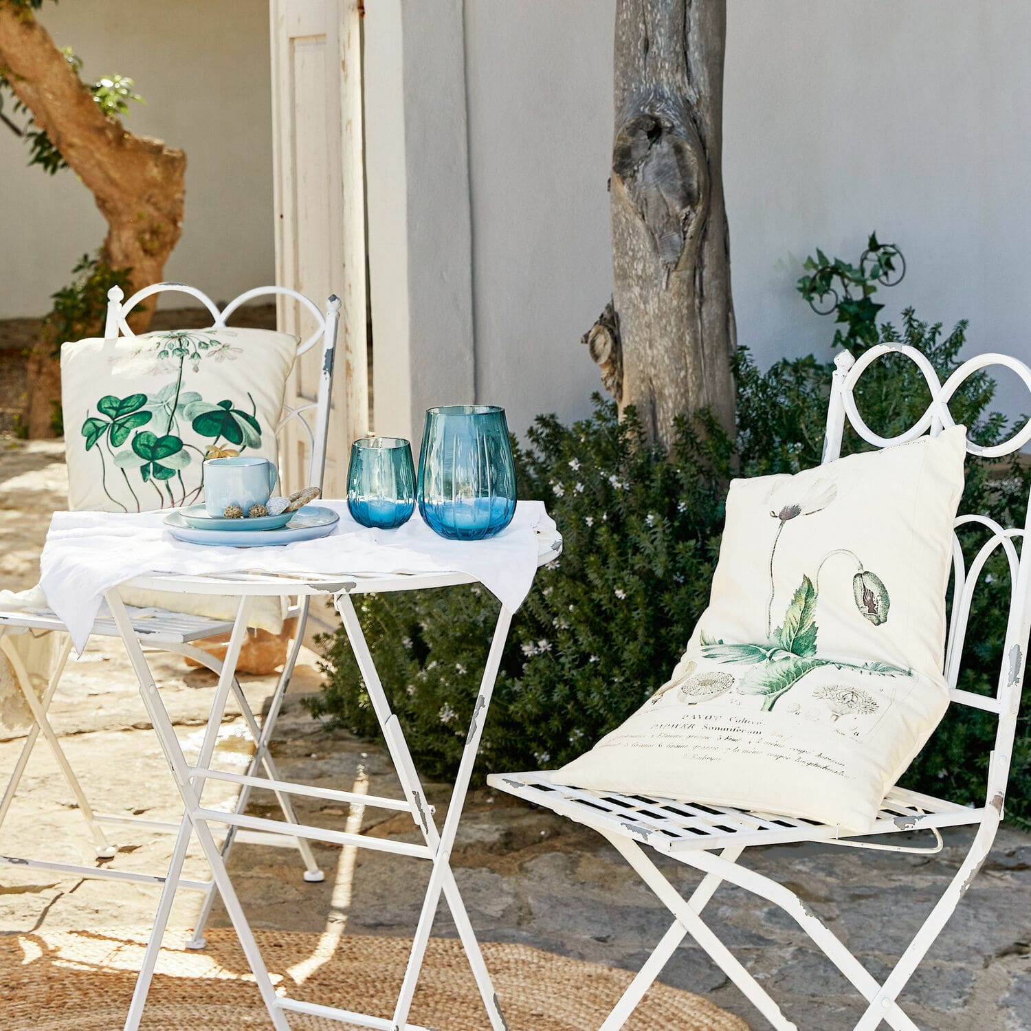 Weißer Metallklapptisch und Stühle mit grünen Pflanzenkissen und blauen Gläsern auf sonniger Terrasse